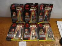 Photo showing six Star Wars Episode 1 action figures in original blister card packaging arranged on a wooden surface.