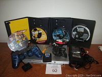 Front view showing the silver PlayStation 2 Slim console, blue translucent Sony wired controller, power supply with cables, 16MB memory card, RCA AV cable, and four game discs with cases arranged in the background.