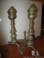 Photo showing two vintage fireplace andirons made of brass and cast iron, positioned side by side showing front and side profiles.