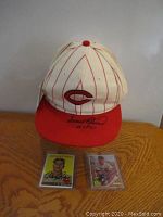 Front view of white and red pinstriped Cincinnati Reds ball cap autographed by Frank Robinson placed behind two vintage signed baseball cards on wood surface.