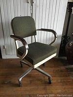 Full view of vintage office chair showing olive green fabric, metal armrests and swivel base with casters on wooden floor next to white paneled wall.