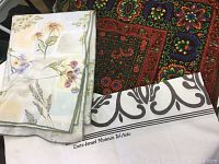 Three tablecloths shown together: white floral design, dark traditional pattern, and white with scroll design and museum text visible.