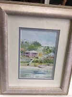 Framed watercolour painting showing a sailboat on the water in front of houses and greenery, with silver frame and matting
