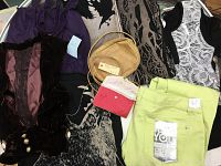 Display of various women's clothing items including tops, a vest, printed dress/top, jeans, and two accessories: a woven tan purse and a small folded item.