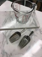 Glass measuring cup with red Fire-King and measurement details, placed on faux marble board with two metal scoops in front