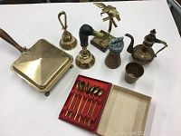 Overview of all 8 brass items on white surface showing a square trivet box, two bells, a pitcher, a small cup, cezve, and the teaspoon set.