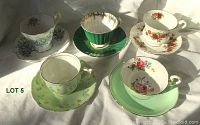 Overview photo of all five vintage china cups and saucers showing floral patterns and gold trim on various base colors including white, green, and cream.