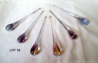 Full view of six solid glass droplets arranged in a semi-circle on white fabric background. Each droplet is long and tapered, displaying an iridescent mauve purple finish.