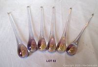 Six heavy glass droplets arranged in a row on a white cloth background. The droplets are long teardrop shapes with an iridescent aurora borealis finish showing purple and some golden reflections.
