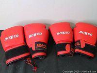 Two pairs of red Boxeo boxing gloves with black wrist straps laid out side by side on a grey floor.