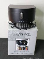 Full view of the black Ash Creek Tie-Matic tie rack on top of its original box showing branding and tie hooks.