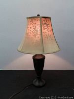 Full view of lamp lit, showing dark wood base and beige fabric shade with floral scroll patterns.
