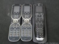 Two identical black and silver universal remotes with display screens and multiple buttons, alongside one black Logitech remote control, all laid out on a black surface.
