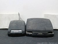 Front view of two black Sharp mini system speakers, one smaller and one larger, both with front silver Sharp labels, surfaces dusty and worn.