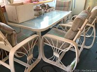 Photo showing rectangular resin framed glass top patio table surrounded by four resin framed rocking chairs with multicolored striped cushions.