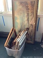 Large landscape painting leaning against wall with plastic bin of framed prints