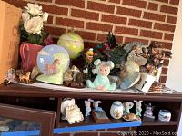 Wide view of multiple figurines including horses, bunnies, clowns, and child bust on wooden furniture against brick wall
