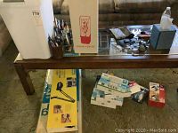 Wide view of lot showing table with pens, pencils in cups, light bulbs, trash bin, and boxes under table including magnifier lamp box and light bulb packages.