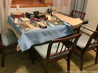 Dining table set covered with cloth on table, four chairs around it, miscellaneous items on table, showing arrangement and context.