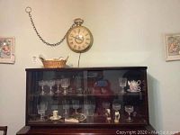 Large wall clock above dark wooden glass-door cabinet filled with various glassware.
