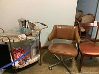 Photo of vintage beverage cart on wheels with assorted glassware and kitchen items, alongside two vintage chairs.