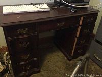 Full view of the wooden desk showing seven drawers with brass handles and various cords and devices on top