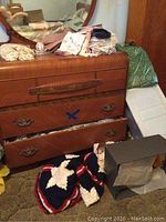 Dresser with piles of folded embroidered and crocheted linens, assorted textiles on top and inside drawer, folded crocheted star-patterned throw and a box on the floor