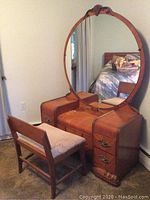Waterfall style wooden vanity with large round mirror mounted, ornate drawer handles, padded center section on top surface, showing wear consistent with vintage use.