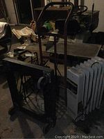 DeLonghi radiator style space heater beside a hand truck, furniture dolly, and small table on wheels in a dimly lit room.