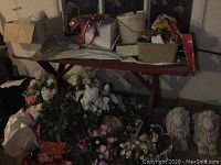 Photo of assorted faux flowers and holiday decoration items piled together with storage containers and other items on table behind.