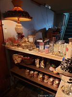 Wide view of a table and shelves holding various mid-century glassware, barware, Baltimore Colts tumblers, an ornate 34-inch lamp with a fabric shade, decorative vases, and ceramic jars on lower shelves. Items are dusty and untested.