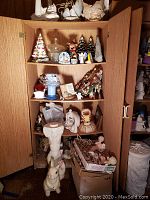 Wooden cupboard with open doors containing multiple shelves filled with assorted figurines and a box of additional figures below.