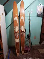 Pair of vintage wood Cascade skis with bindings, fishing poles, and badminton racket side by side against wall