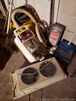Holmes dual window fan, Montgomery Ward canister vacuum, Genie yellow wet/dry vacuum, Bissell box, blue dustpan and brush, all showing wear and dust.