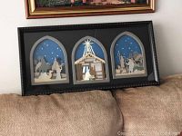 Full view of the framed nativity shadow box showing three arched panels with paper cut designs against a blue background.