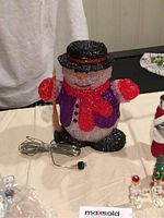 Glittery acrylic snowman decoration with black hat, red scarf, broom, and electric cord shown on table
