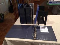 Two 3-inch ring binders and one black plastic file holder on a table, binders standing upright with one slightly open.