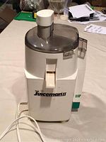 Front and side view of white and gray plastic electric juicer with 'The Juiceman' branding, power cord visible, transparent top lid, chute for feeding fruit, spout for dispensing juice.