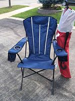 Two blue folding outdoor chairs set on a driveway, each with a cup holder on the armrest and a red and white carrying bag standing upright beside the chairs.