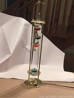 Full view of the Galileo thermometer with colorful floating spheres inside a tall glass tube supported by brass-colored accents.