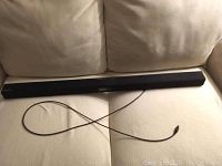 Front view of Samsung black sound bar laid on beige couch with attached cable visible.