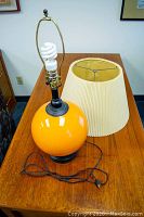 Orange glass base table lamp with attached lightbulb socket and cord, placed on a wooden table, no shade attached.