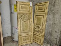 Two tall vintage wooden shutters leaning against a wall, showing the carved floral medallion panels and rectangular recessed panels with detailed molding trim. Painted cream color with signs of use and age.