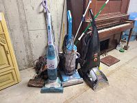 Photo shows a row of upright vacuums and a steam mop resting against a concrete wall with a piano visible in background.