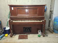 Full front view of the upright wooden Marshall and Wendell player piano in a basement or garage setting showing overall condition and player piano mechanism panel near the bottom.