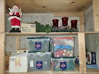 Two wooden shelves containing Christmas decor items including Santa figurine, garland, red candle holders, plastic storage bins with ornaments and other holiday decorations.