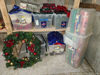 Photo showing stacked clear plastic storage boxes filled with assorted Christmas decorations, wreath on floor with red bows and gold/red ornaments.