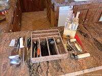 Wide shot of 39-piece black matte flatware set arranged in woven flatware organizer tray alongside Sabatier knife block and various kitchen utensils on granite kitchen counter