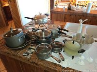 Overview of 14 pots and pans including Calphalon and Pampered Chef pieces arranged on countertop, showing varying sizes and a fondue set.