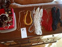 Assorted costume jewelry laid out, including multi-strand beaded necklaces in black, red, white, tan, chain necklace, and a red container with additional jewelry pieces including bracelets, earrings, and watches.